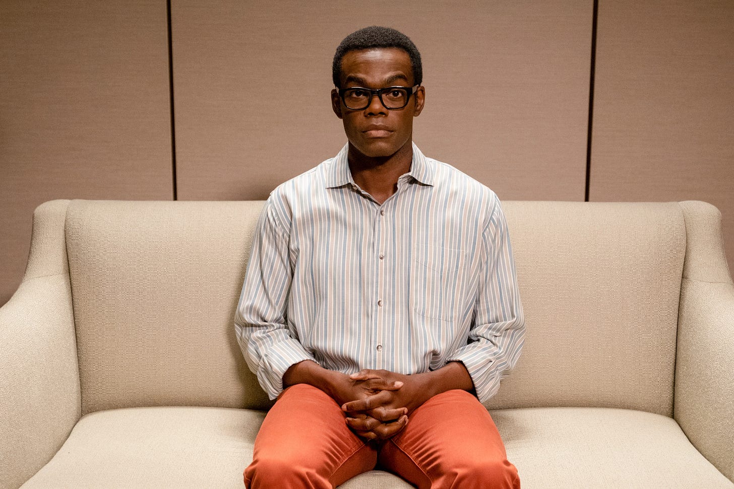 Chidi Anagonye, a Black man with glasses in a striped shirt, sits on a couch