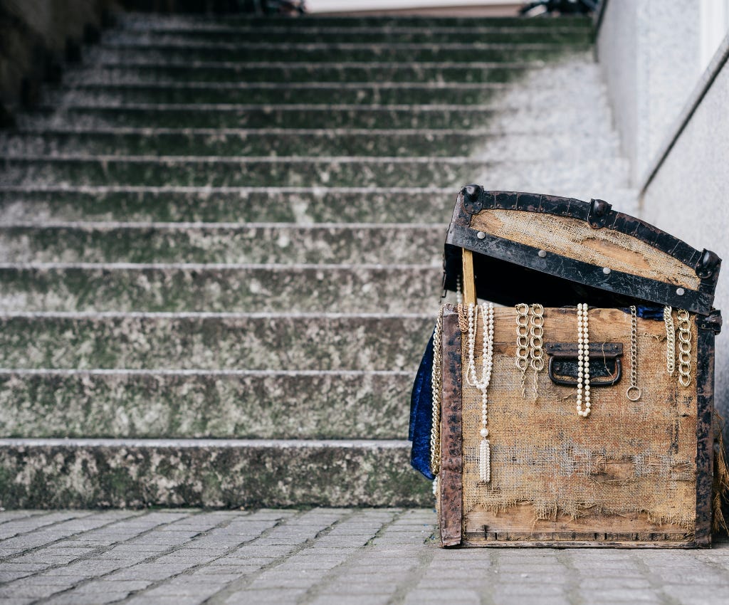 Old battered faded brown sea treasure chest with black flat iron around the partially opened lid.  Hanging over the side of the treasure chest at the lid opening are several pieces of jewelry. Six are gold necklaces in varying designs and lengths. Two of them are strings of pearls. The chest is sitting on grey brick pavement.  To the left of the chest is a steep stone marble colored stairway of dark grey, green and grayish white.