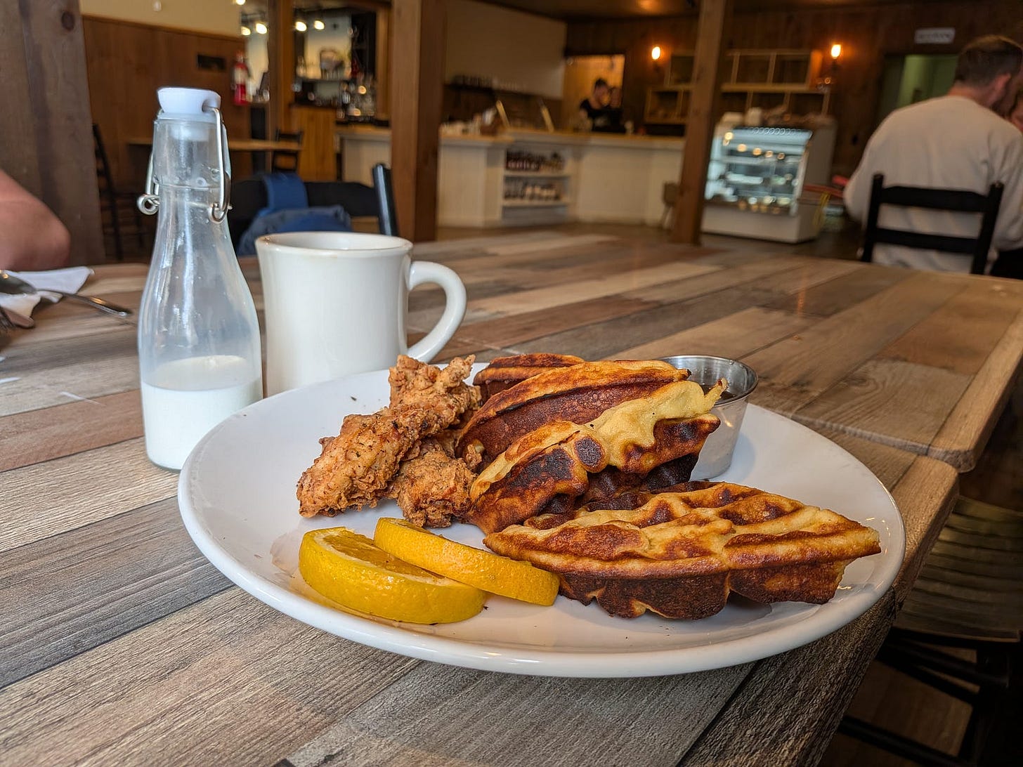 A plate of chicken and waffles on a rustic wooden table at a Vermont cafe. Golden-brown waffles are stacked alongside crispy fried chicken, garnished with orange slices and a small cup of maple syrup. A glass bottle of fresh local milk and a white coffee mug sit beside the plate, with the warm, wood-paneled restaurant interior visible in the background. A plate of chicken and waffles on a rustic wooden table at a Vermont cafe. Golden-brown waffles are stacked alongside crispy fried chicken, garnished with orange slices and a small cup of maple syrup. A glass bottle of fresh local milk and a white coffee mug sit beside the plate, with the warm, wood-paneled restaurant interior visible in the background.