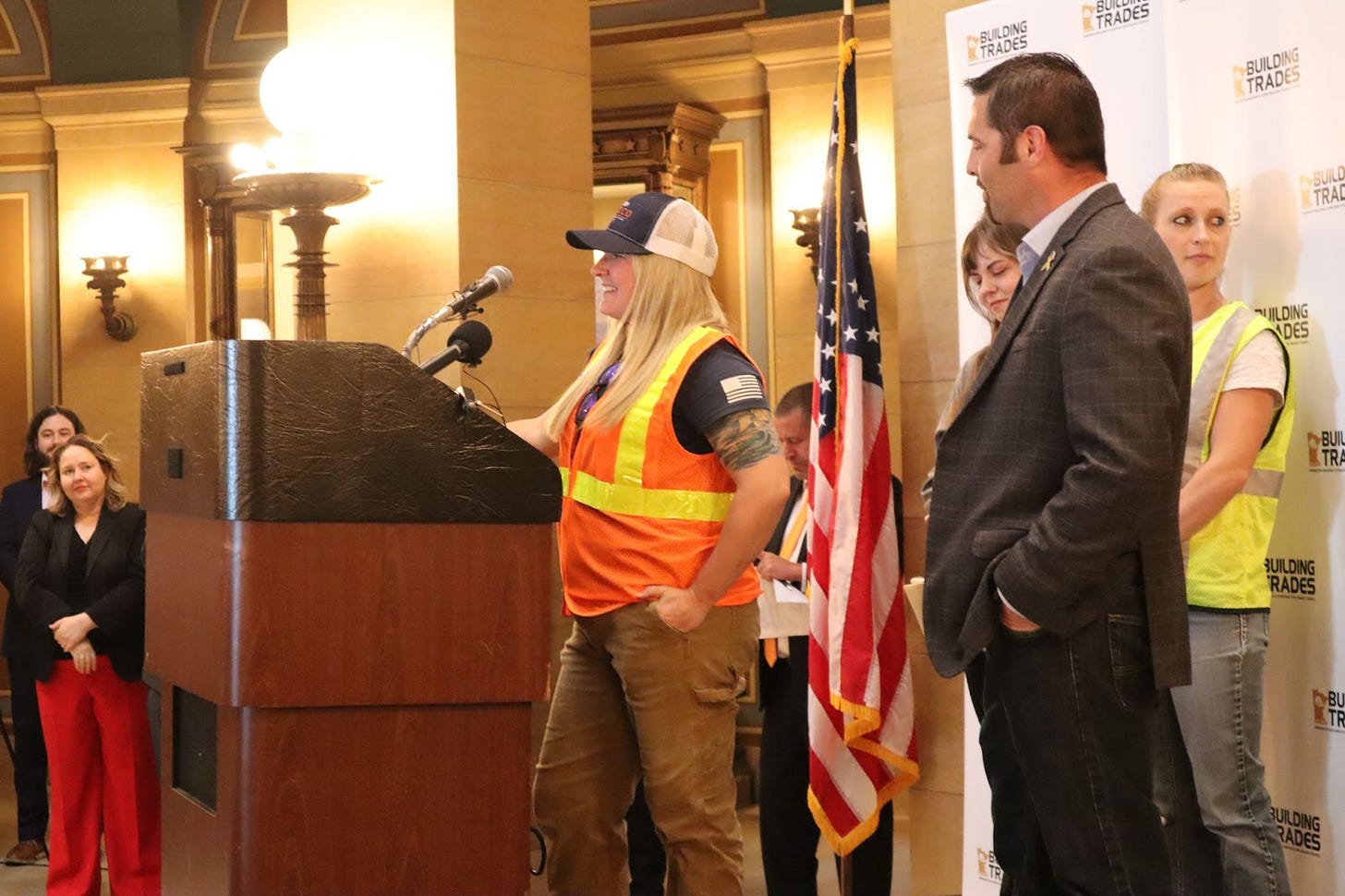 a blonde woman wearing a cap stands behind a podium wearing an orange construction vest, a man in a grey suit stands behind her a blonde woman wearing a cap stands behind a podium wearing an orange construction vest, a man in a grey suit stands behind her