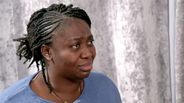 A lady with a light blue top and dark hair tied back in corn rows speaks to the reporter, with light grey curtains in the background A lady with a light blue top and dark hair tied back in corn rows speaks to the reporter, with light grey curtains in the background