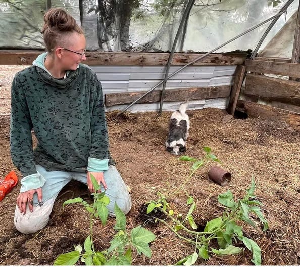 A nonbinary person kneeling in a greenhouse among plants, looking at a small black and white dog and smiling.