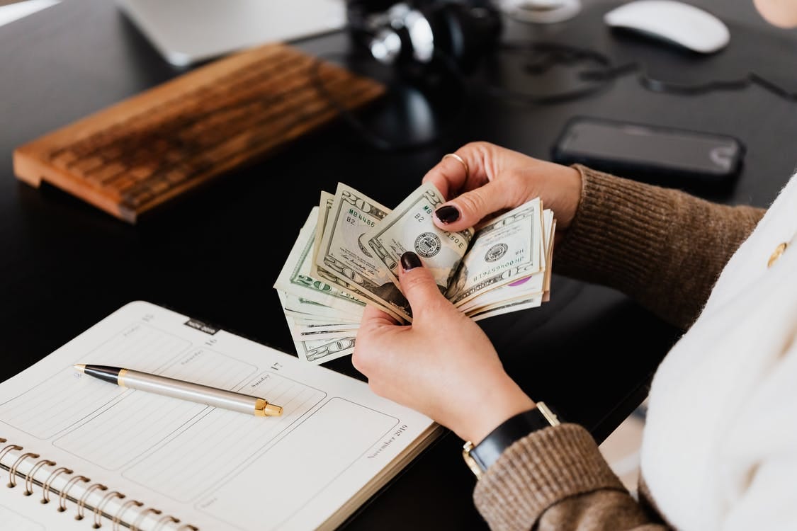 Unrecognizable elegant female in sweater counting dollar bills while sitting at wooden table with planner and pen Unrecognizable elegant female in sweater counting dollar bills while sitting at wooden table with planner and pen