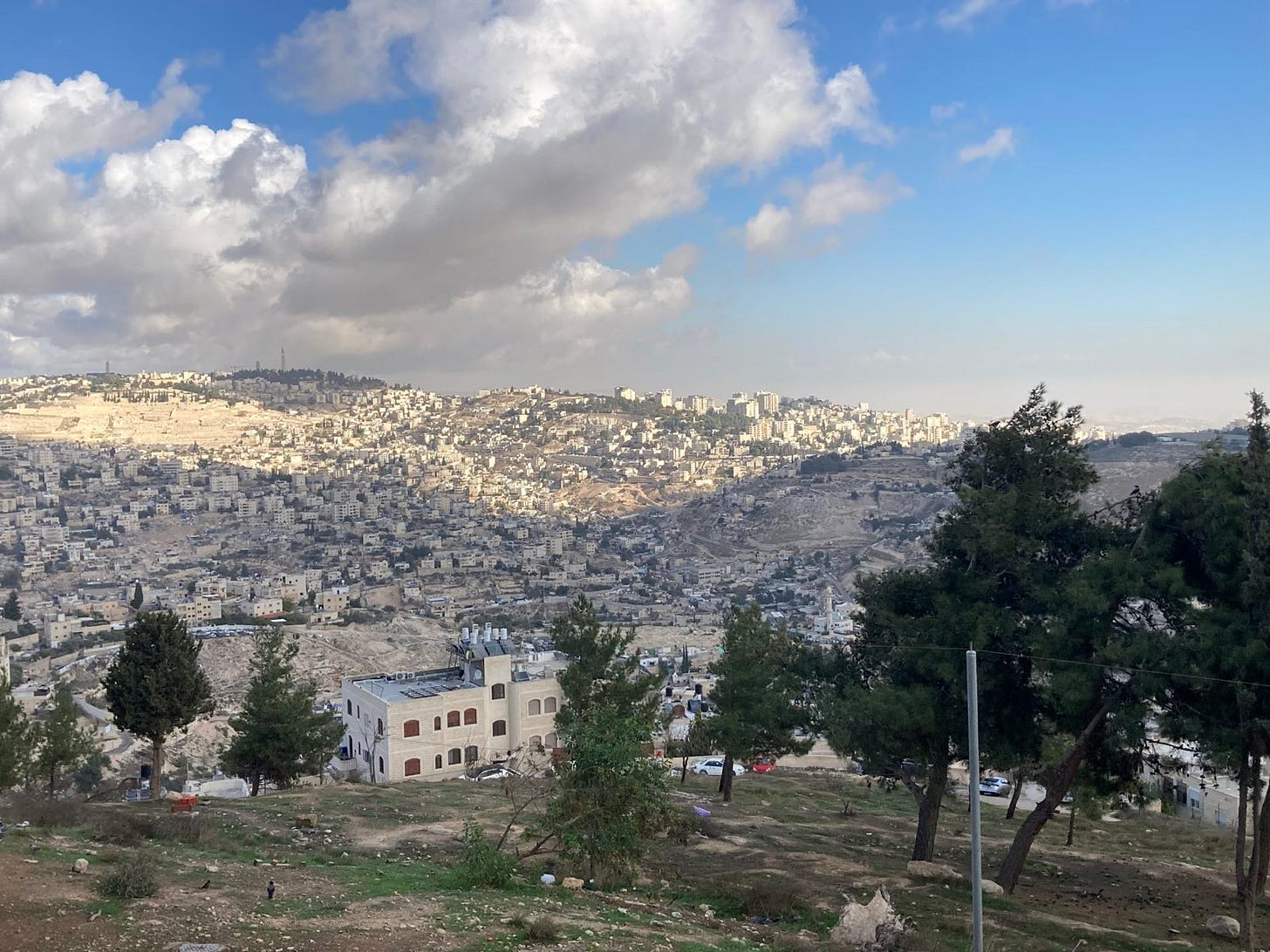 A photo taken from a hilltop looking out over Jerusalem and surrounding areas. A cloudy sky over miles of beige multistory buildings. A photo taken from a hilltop looking out over Jerusalem and surrounding areas. A cloudy sky over miles of beige multistory buildings.