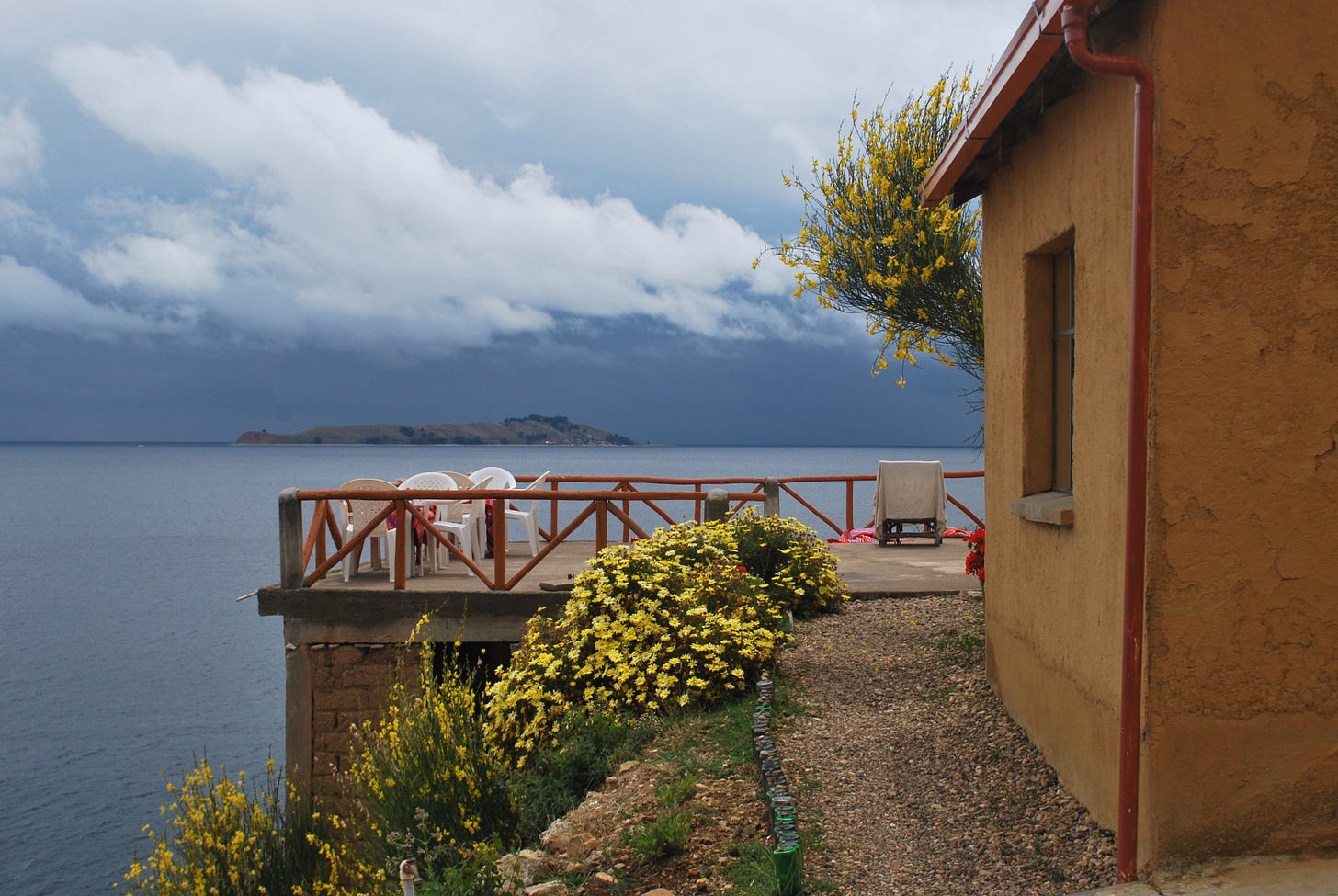 The photo shows the island Isla del Sol on the Lake Titicaca in Bolivia. To the right, there is a sand-coloured house, there is a terrace overlooking the lake, yellow flowers in the lefthand corner. Some low-hanging clouds are visible and it is unclear whether the weather will hold or change. The photo shows the island Isla del Sol on the Lake Titicaca in Bolivia. To the right, there is a sand-coloured house, there is a terrace overlooking the lake, yellow flowers in the lefthand corner. Some low-hanging clouds are visible and it is unclear whether the weather will hold or change.