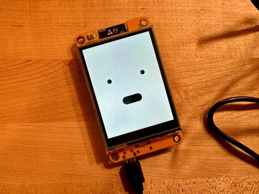 Photo of a small yellow ESP32 display on a wooden desk, showing a simple cartoon face with two eyes and an open mouth on a white screen.