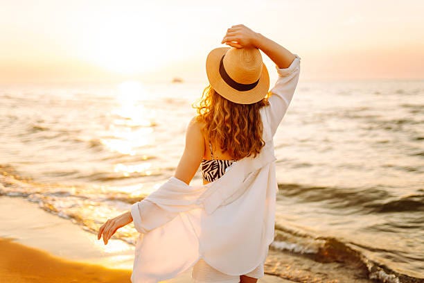 A happy woman is having fun on the beach in the sun's rays. A happy woman is having fun on the beach in the sun's rays. A beautiful woman in a hat smiles and enjoys the sunset by the sea. The concept of relaxation, freedom, and enjoyment. A Beauty by the Sea stock pictures, royalty-free photos & images
