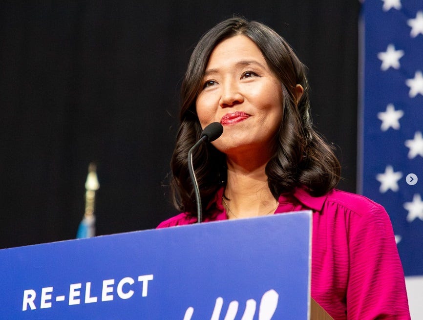 Michelle Wu stands at a lectern with 'Re-elect Michelle Wu!' written on it, in front of an American flag backdrop. 