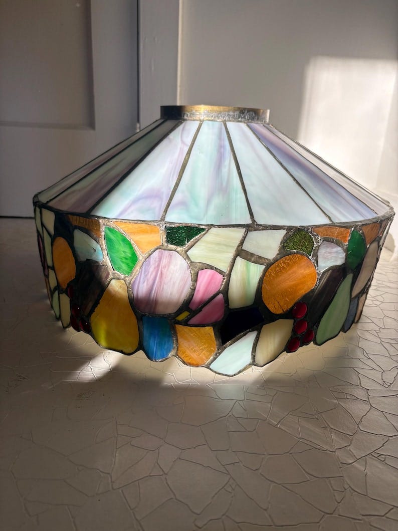May include: A stained glass lamp shade with a colorful mosaic design. The shade features a variety of colors, including blue, green, orange, pink, and white. The top of the shade is a light blue and purple color. The shade has a brass-colored metal rim.