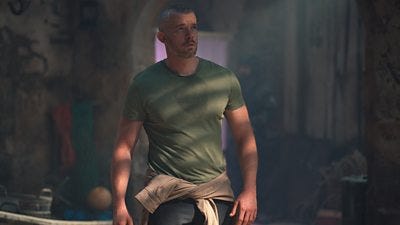 A man in a t-shirt stands in the shadows looking up at something off screen