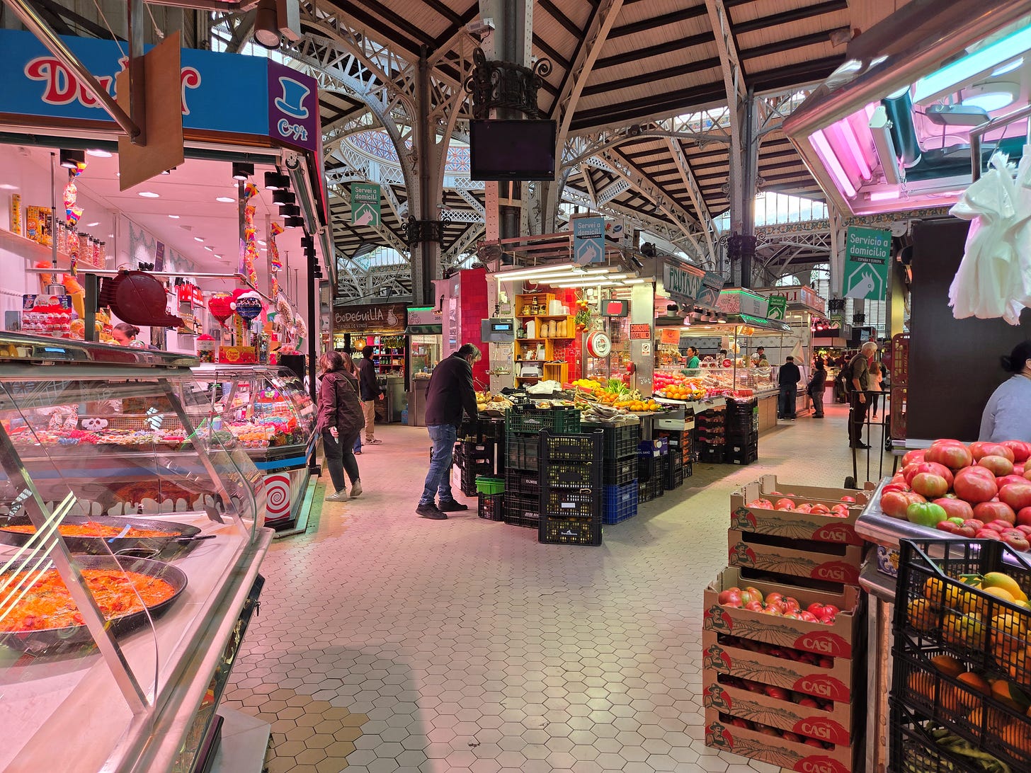 A spacious indoor market with high arched ceilings, bright food stalls, crates of fresh produce, pastries, and shoppers walking through the aisles under warm pink and natural light.
