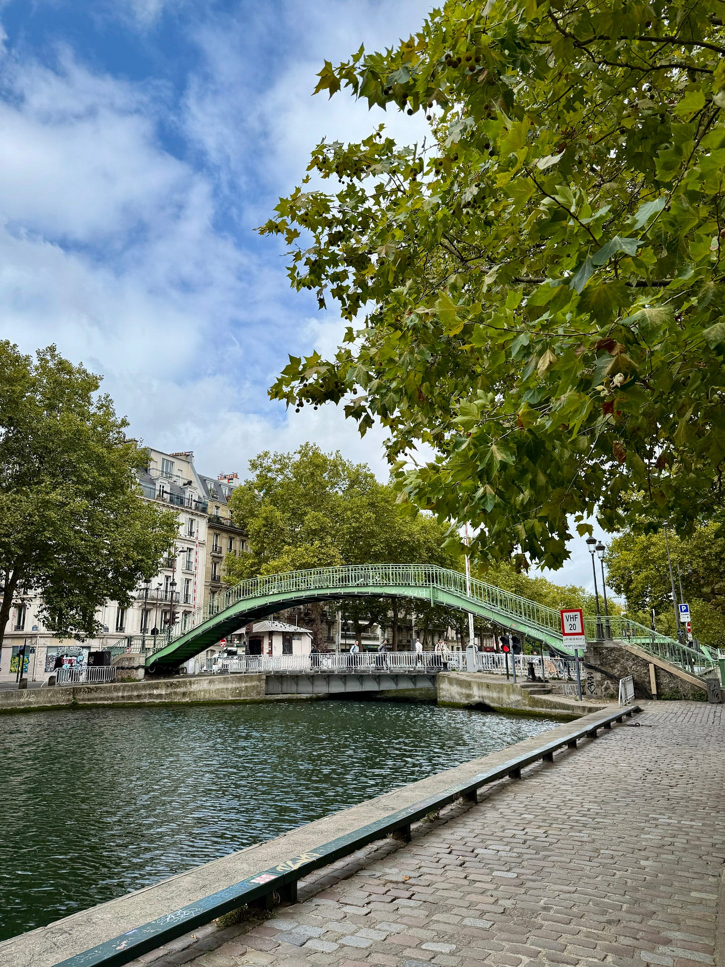Canal Saint-Martin in Paris on an overcast day, with a green iron footbridge spanning the water and cobbled walkways lining the canal.