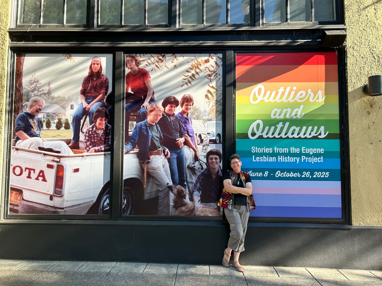 A storefront window display features historic photographs of a group of people seated in and around a pickup truck. To the right, a large rainbow‑colored poster reads “Outliers and Outlaws: Stories from the Eugene Lesbian History Project” with dates “June 8 – October 26, 2025.” In front of the window, one person stands with arms crossed, leaning against the display.