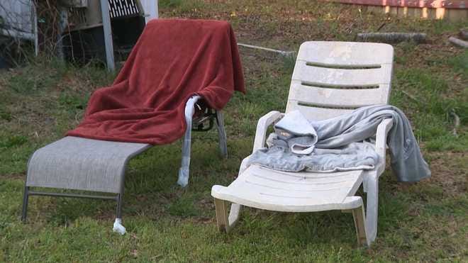 Two weathered lawn chairs sit on a grass yard — one metal with a red towel draped over it, one white plastic with clothing left on the seat.