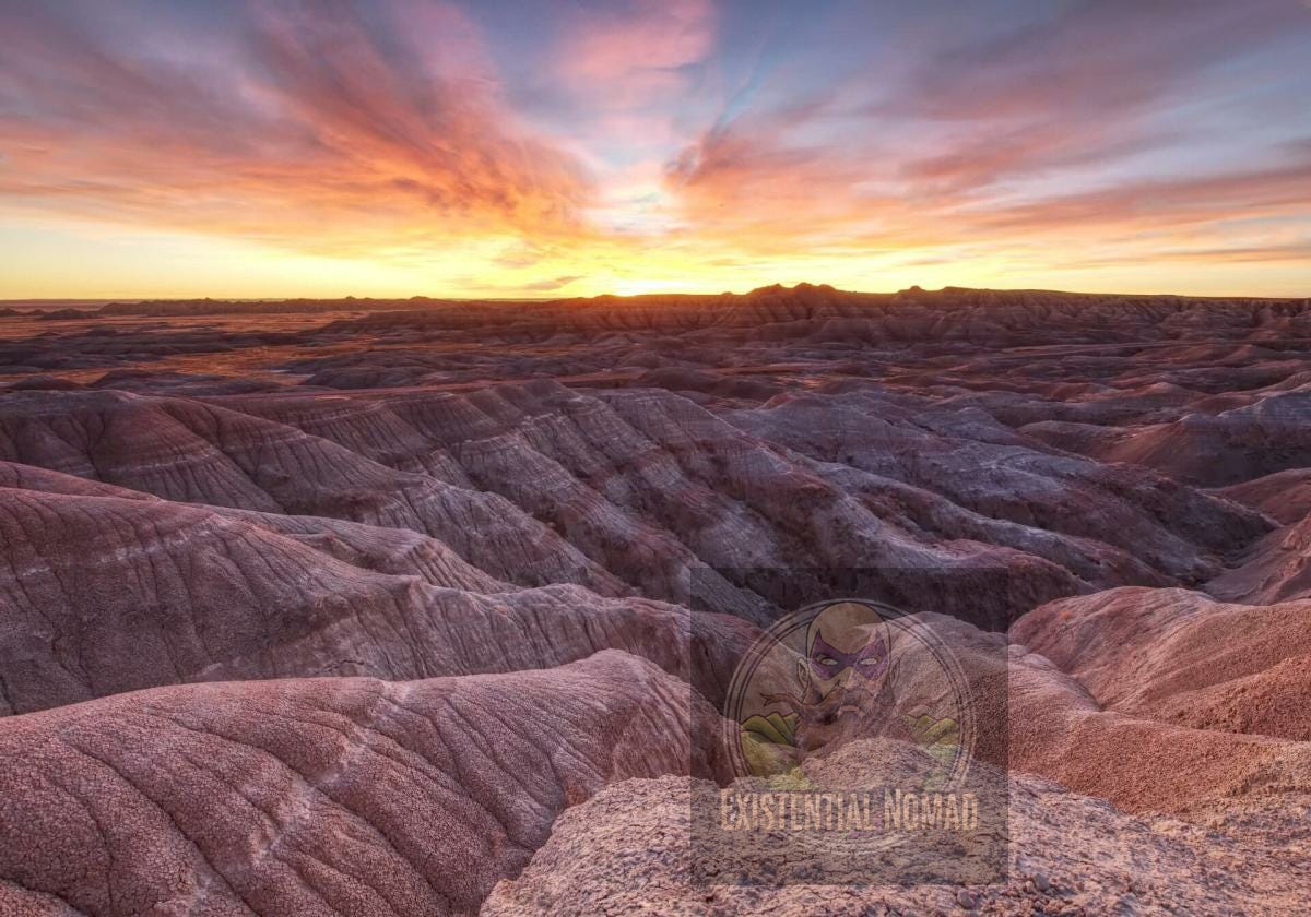 This is a landscape photograph of a rugged, arid terrain, likely the Badlands, at sunrise or sunset. The ground is composed of rolling hills and ridges with deep crevasses and a mix of soft, muted colors, including purples, pinks, and browns. In the background, the sun is setting (or rising) over the horizon, casting a warm, golden glow and creating a dramatic sky filled with vibrant orange, pink, and yellow clouds. The overall mood of the image is serene and majestic.
