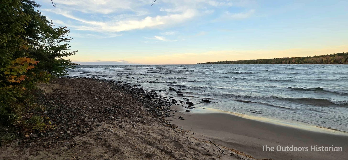 A sandy shore transitions to small and medium sized cobble. The lake is wavy and the sky is blue with a soft glow at the horizon.