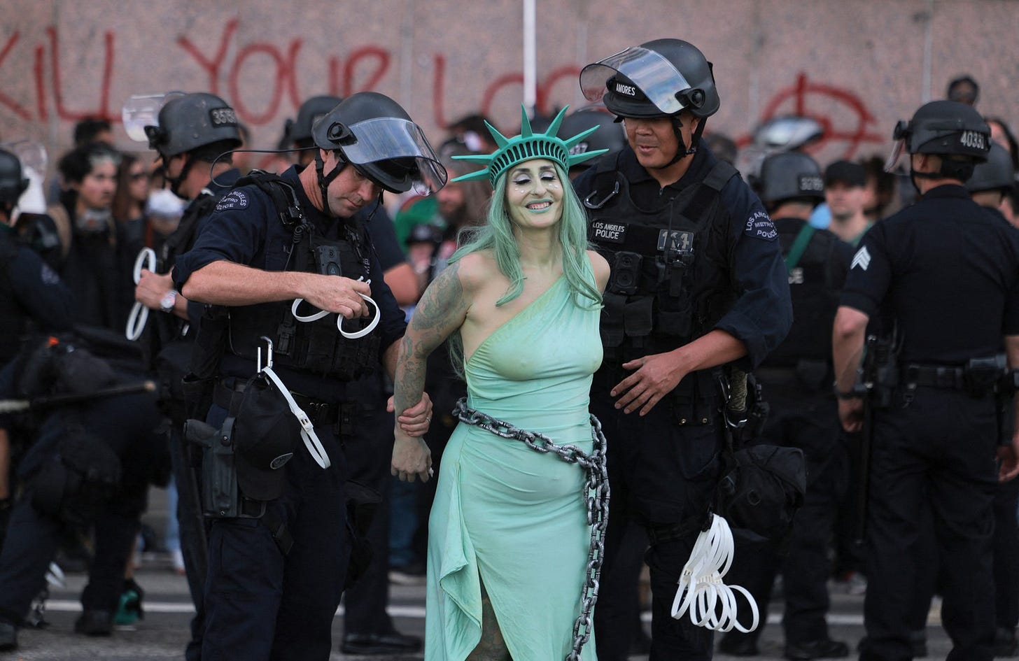 Police officers in riot gear arrest a protester dressed as Lady Liberty in chains.