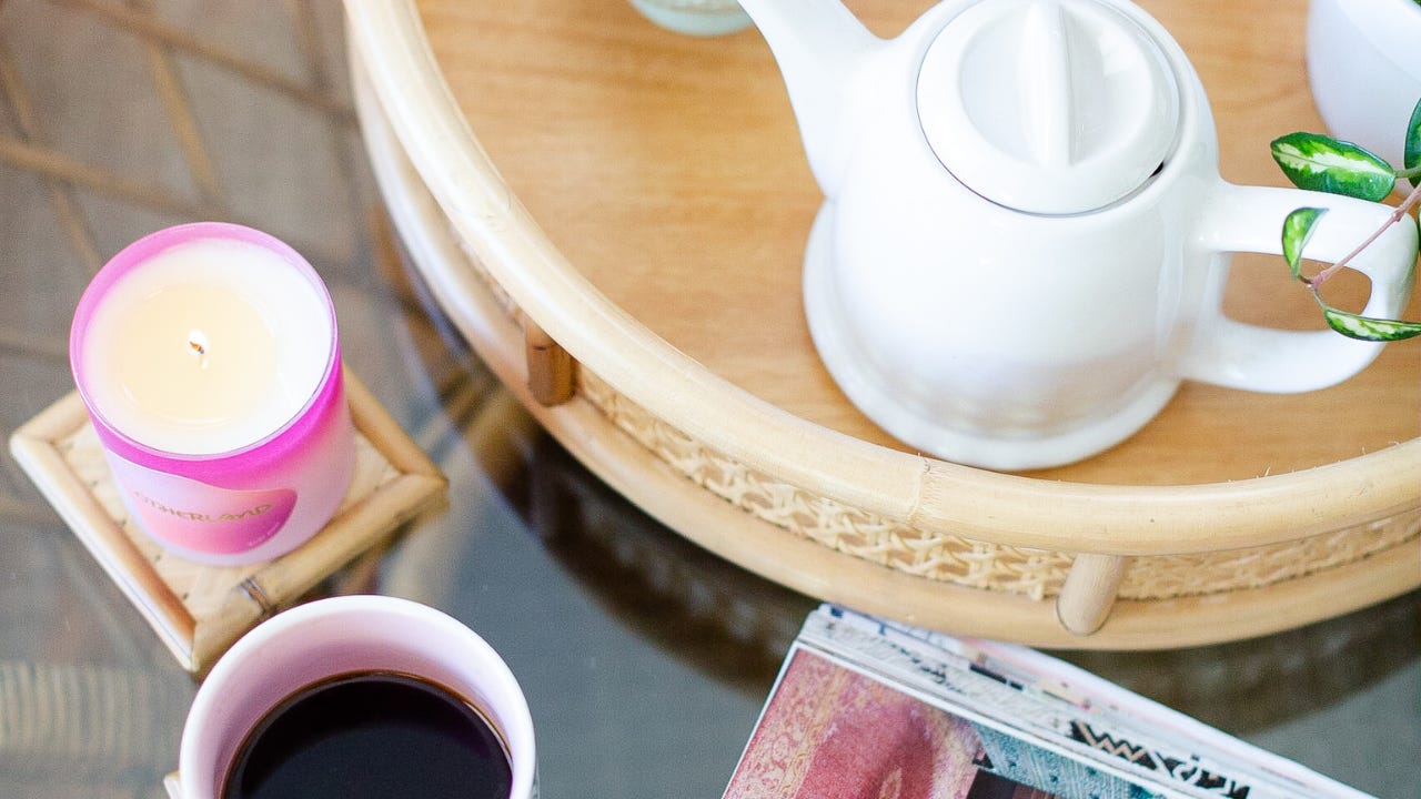 A cup of coffee sits on a glass table beside a lit pink candle, a white teapot on a woven tray, and a magazine. A cup of coffee sits on a glass table beside a lit pink candle, a white teapot on a woven tray, and a magazine.