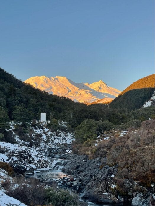 Snow-capped mountain peaks glowing orange at sunrise above rocky alpine stream