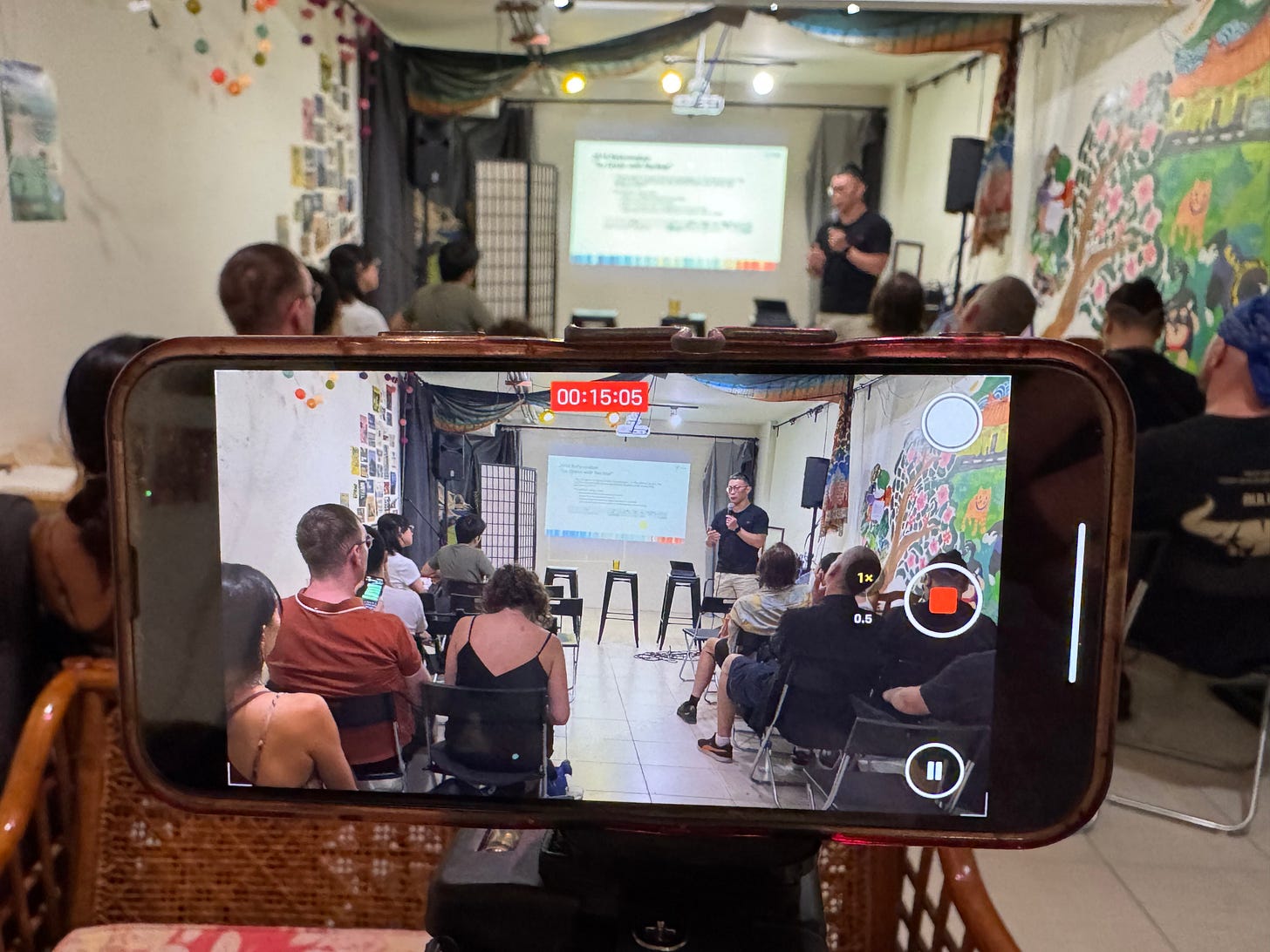 Horizontal photo of a close up of a smartphone on a tripod filming a lecture at the Daybreak basement, with the camera recording view showing the speaker in focus, while the event itself is seen out of focus in the background.