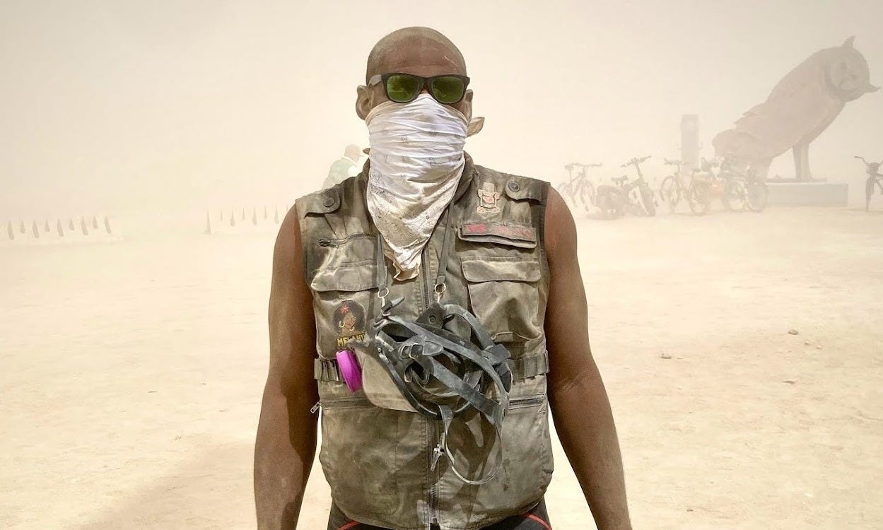 A dusty man wearing a black ranger vest and a white bandana dust mask and sunglasses stands in the middle of a dust storm at Burning Man