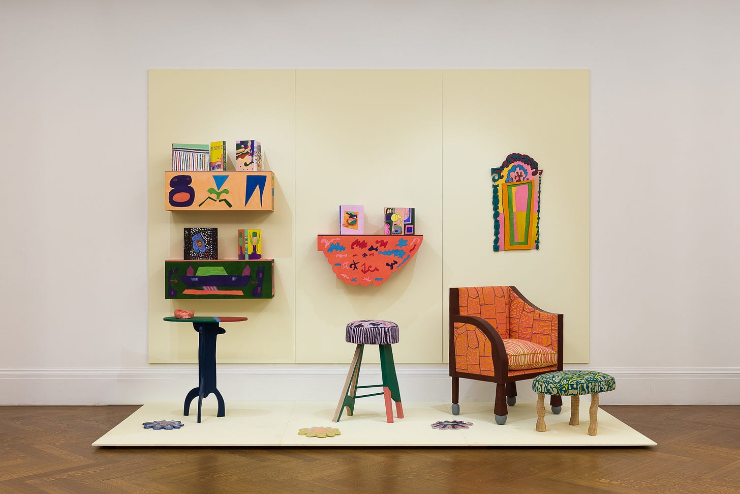 A photograph of a gallery wall containing decorative graphic shelves with books. Infront of the wall are four pieces of unique furniture