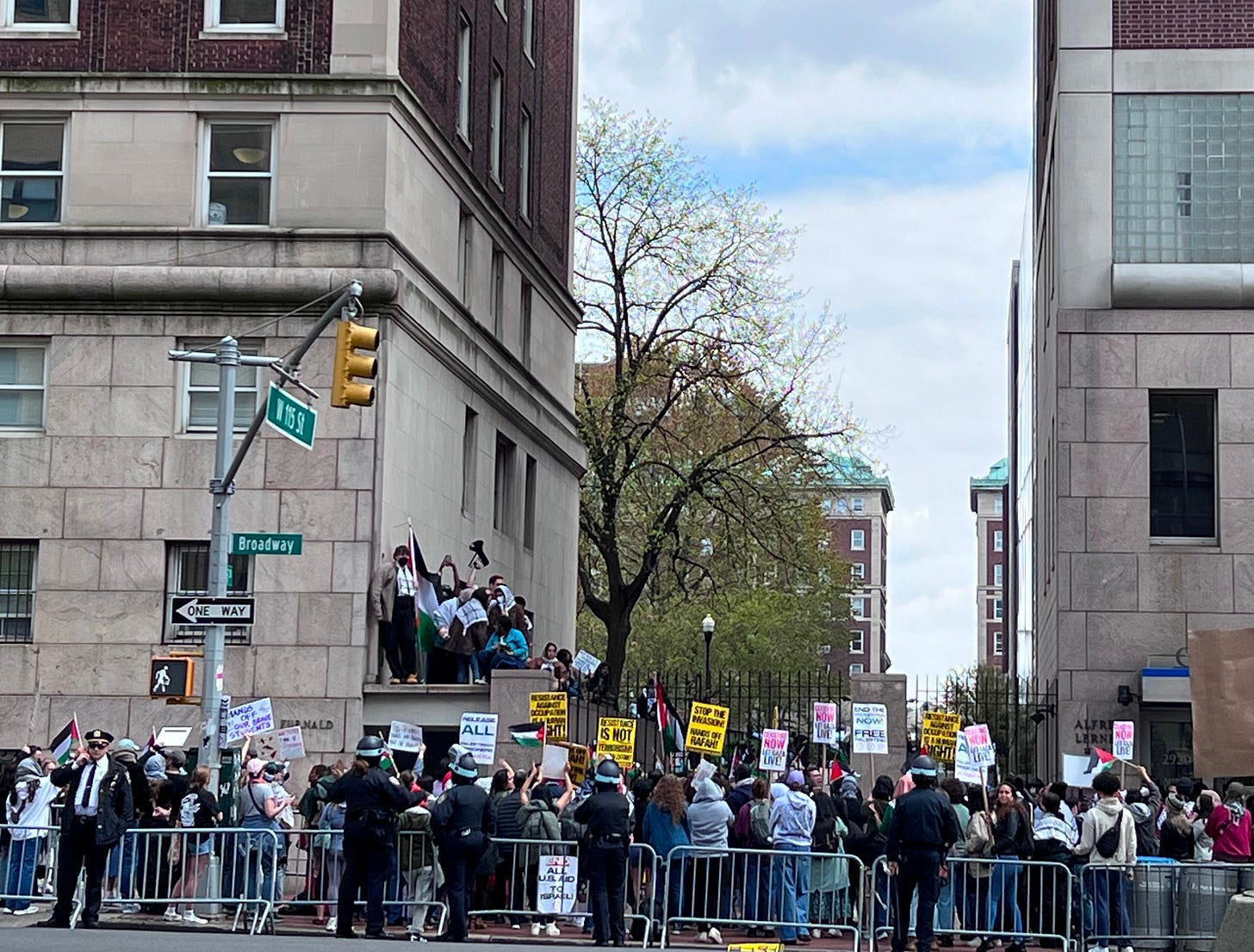 A protest outside the gates of a university; students are carrying signs and there are a few police present.