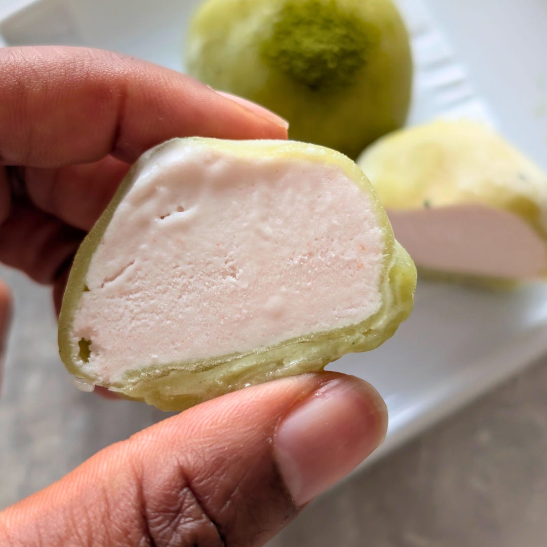 Hand holding a matcha mochi ice cream filled with strawberry ice cream, showing the chewy green wrapper and pink filling.