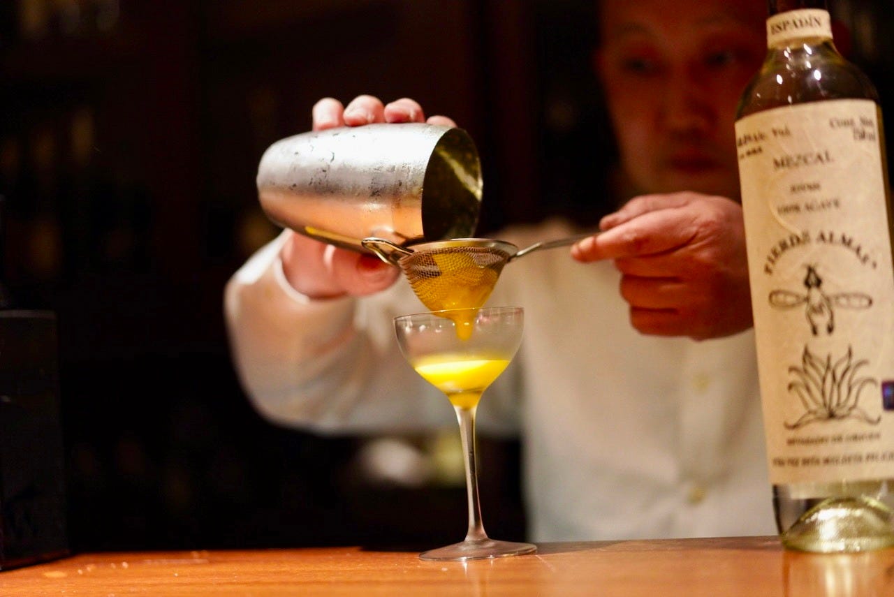 Someone pouring a drink from a metal cup into a glass through a strainer.