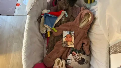 A baby cot is pictured in a room inside Keira's apartment. In the middle of the cot are baby clothes and blankets and photos of Zammi.