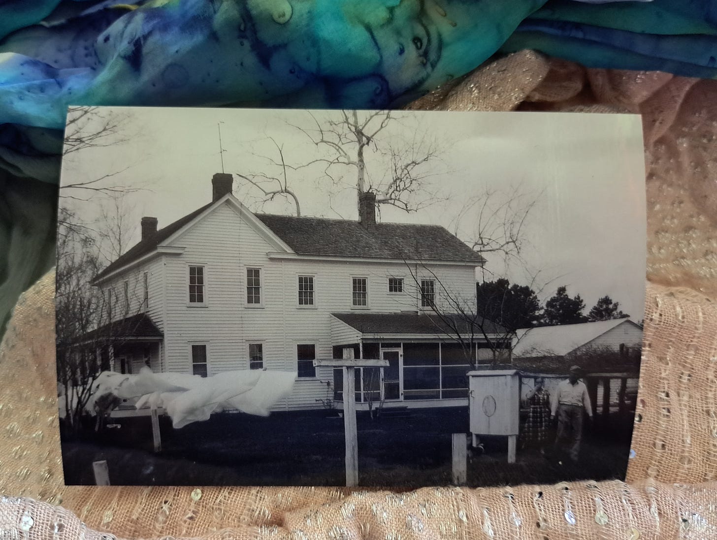 Black-and-white photograph of a white frame house with sheets blowing on a clothesline in front. The photo is positioned on a background of scarves; the top scarf is shades of blue and green, the bottom one is light peach with scattered sequins.