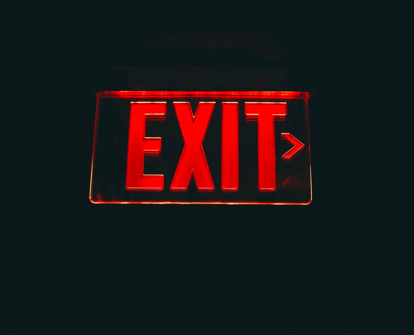 A glass sign lit up with red light illuminating the letters "E-X-I-T" on a dark black background. It almost looks as if the exit sign is imply floating in a black void.