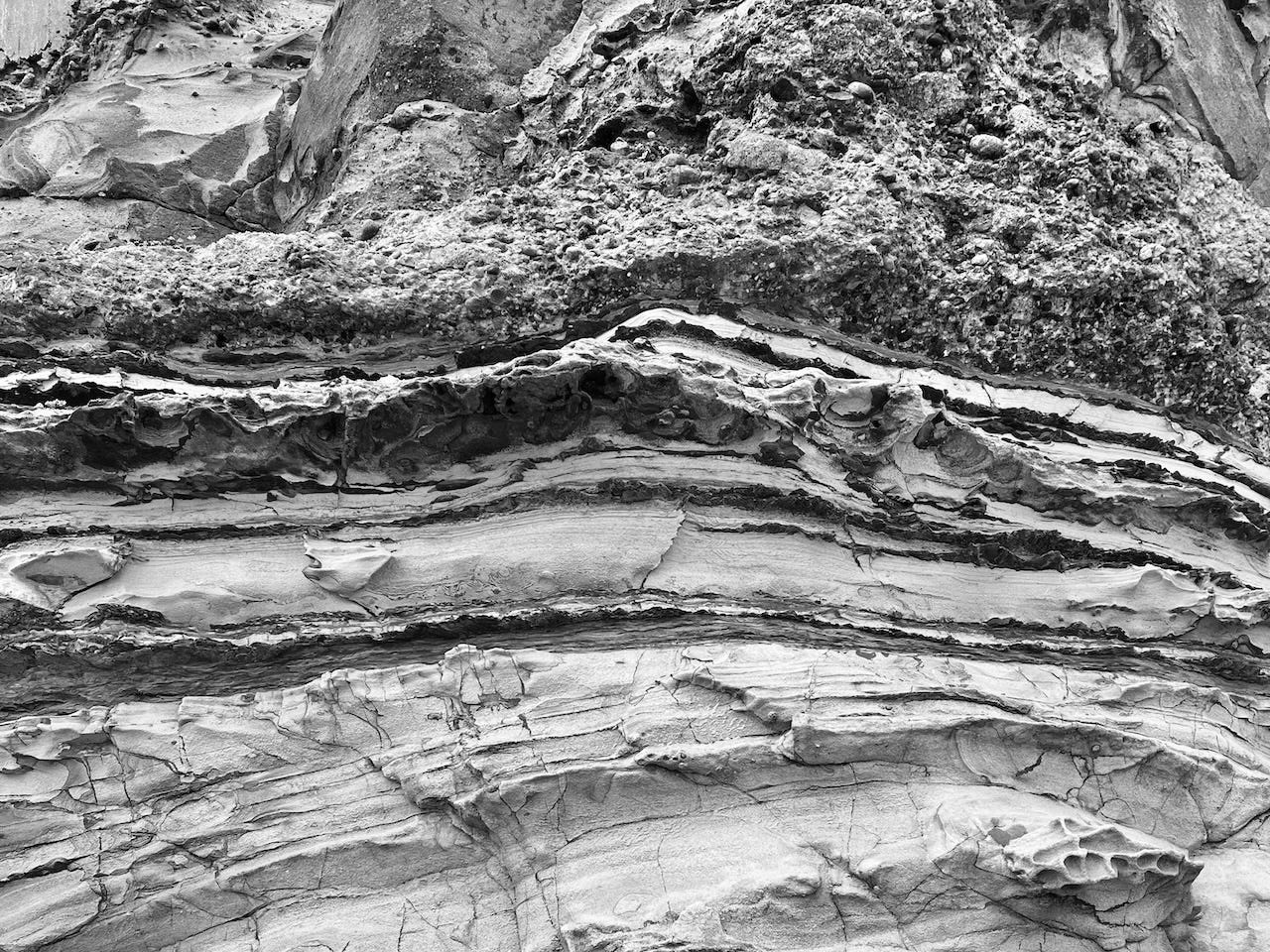 GPT-4o: This is a black-and-white photograph showcasing the intricate layers of sedimentary rock. The image highlights detailed textures, stratification, and patterns formed over time due to geological processes. The contrast emphasizes the distinct lines and weathered surfaces, presenting an artistic perspective on natural rock formations.