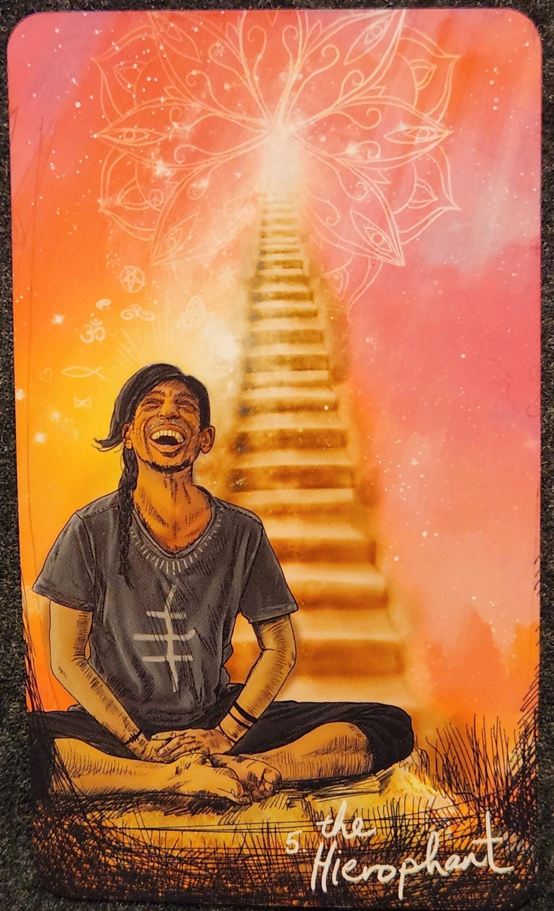 A tarot card labelled 5. The Hierophant. The card shows a man with long hair tied into a ponytail. He is sitting cross legged with his hands resting in his lap. He is smiling with his eyes closed and face upturned. Behind him is a set of stairs leading upwards. A mandala symbol is etched in light in the sky at the top of the stairs. The sky is a brilliant pink. 