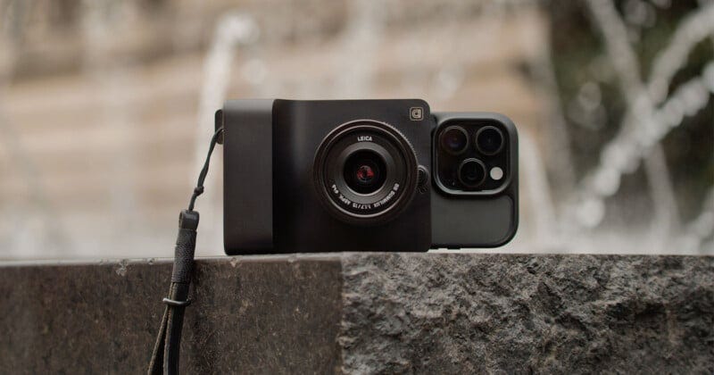 Um smartphone com uma câmera Leica e uma pulseira repousa sobre uma superfície de pedra, com uma fonte desfocada ao fundo.