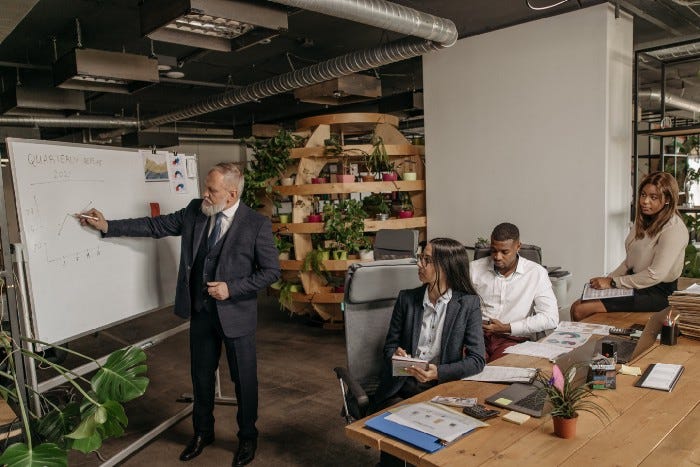 An elderly man in a business suit gesturing towards a white board, with younger colleagues having a mix of emotions. Some are paying attention and taking notes, while others are looking down. An elderly man in a business suit gesturing towards a white board, with younger colleagues having a mix of emotions. Some are paying attention and taking notes, while others are looking down.