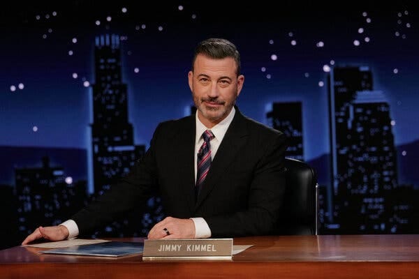 Jimmy Kimmel, in a dark suit, sits at a desk on his studio’s set. Jimmy Kimmel, in a dark suit, sits at a desk on his studio’s set.