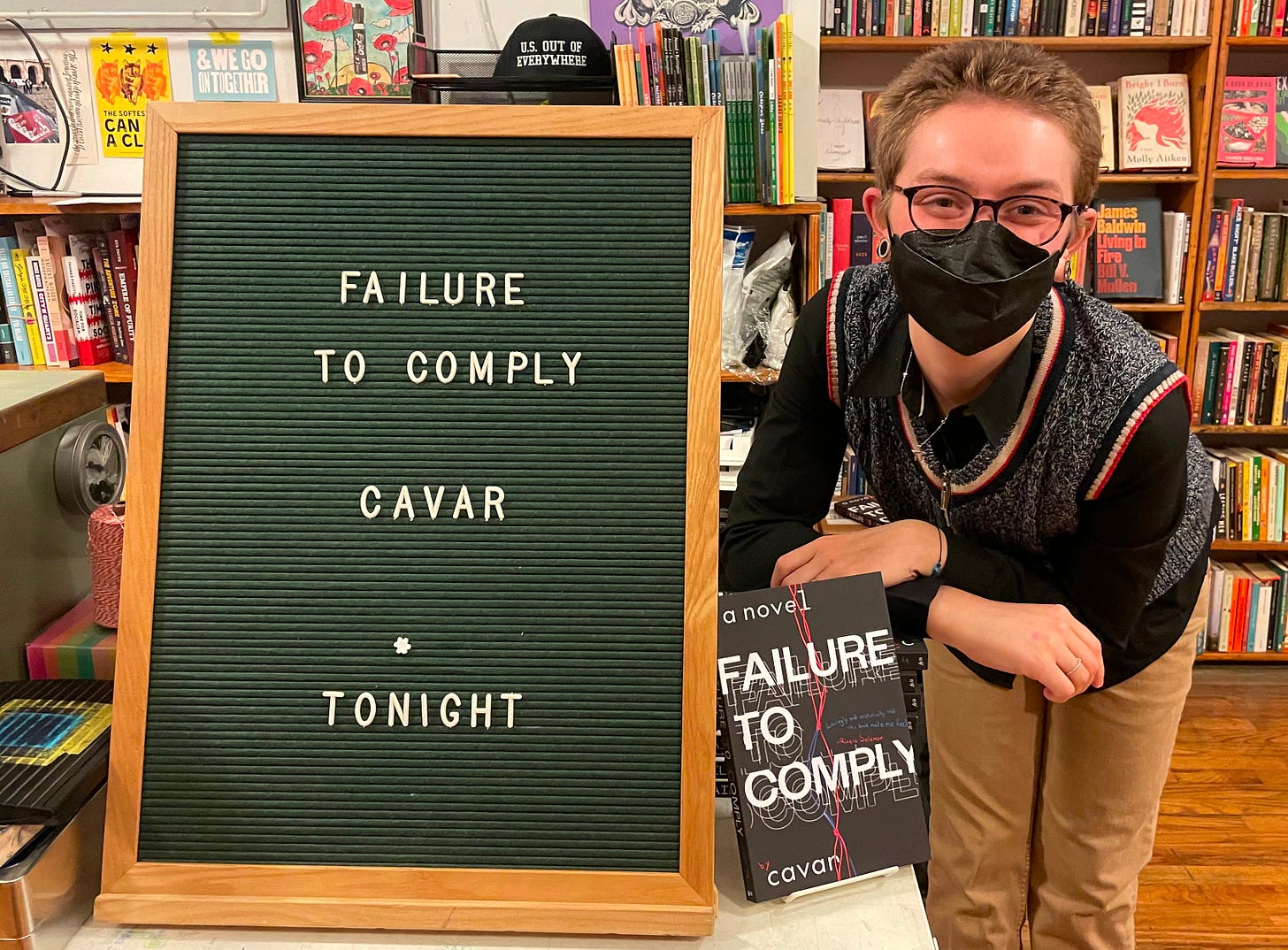 Cavar, a genderless white person with glasses, a black mask, a sweatervest and black turtleneck, and khakis, poses beside a sign announcing their event.