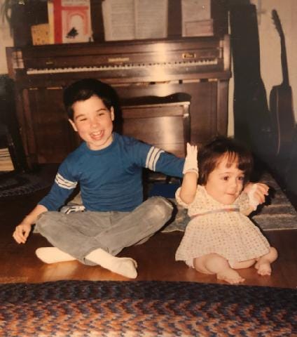 A young boy with dark brown hair is sitting on the ground cross-legged, he is dancing with a very tiny Gaelynn Lea, who is wearing a frilly dress and white gloves and spinning around in a circle on the floor, without a wheelchair in sight.