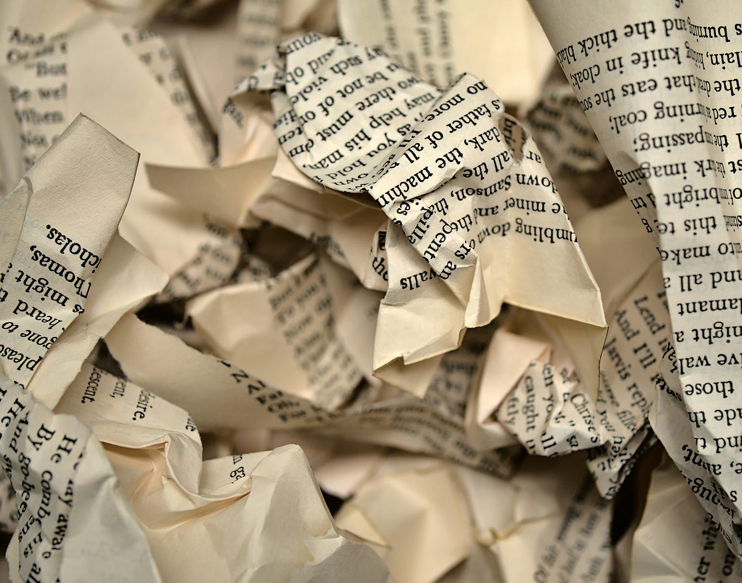 The image shows crumpled pieces of paper with printed text visible on them. Various torn and wadded-up paper fragments are scattered throughout the frame, creating a textured pile of discarded writing or documents. The text appears to be from books or printed materials, though individual words are only partially legible due to the crumpling. This visual could represent writer's block, revision process, discarded ideas, or the frustration of drafting and editing content.
