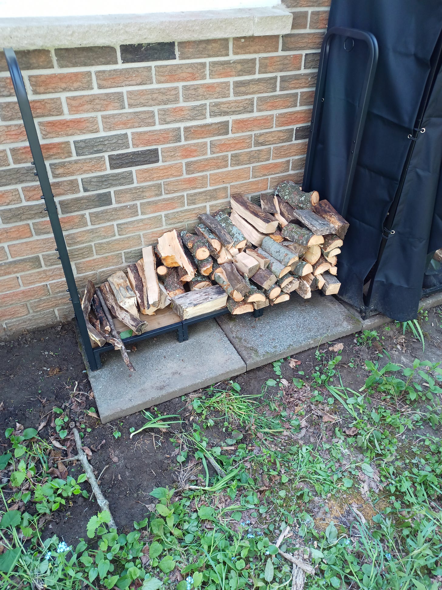 A black steel rack about 120cm wide and 120cm high. The rack is about 15% filled with two rows of wood, with shorter pieces in the front row. Each piece of wood is on average less than 6cm by 6cm when viewed from the end.