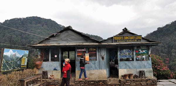 Metal shack sitting on the side of a mountain. There is a woman wearing a red coat in front and grey skies behind. 