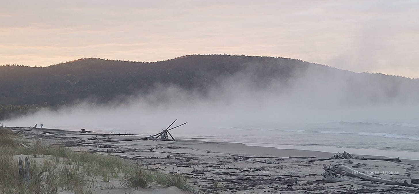 A morning view from the beach at Neys Provincial Park. Wisps of white sea smoke rise off the cold water, framed by the rugged, dark silhouettes of the Canadian Shield topography under a soft morning sun.