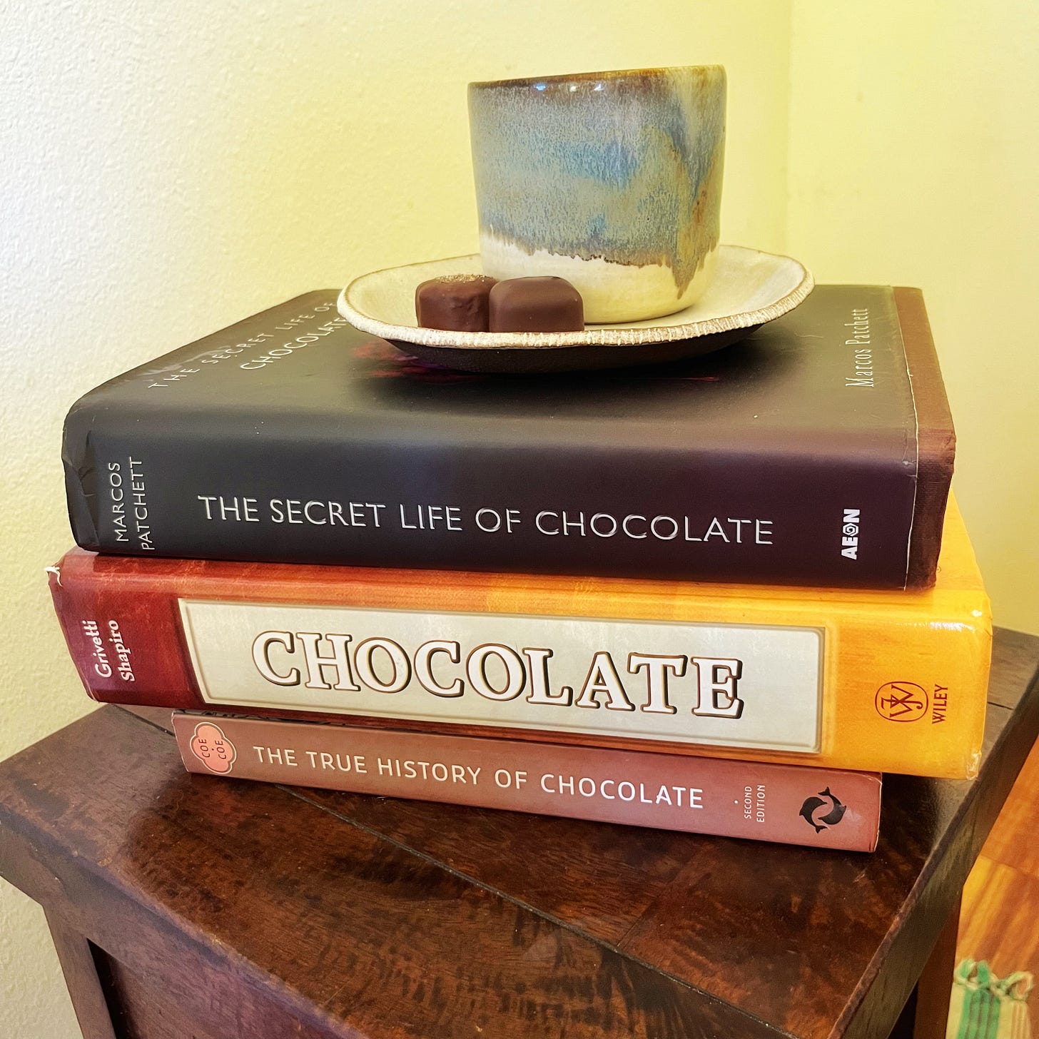 Table stacked with three books, a cup, a saucer, and two chocolates 