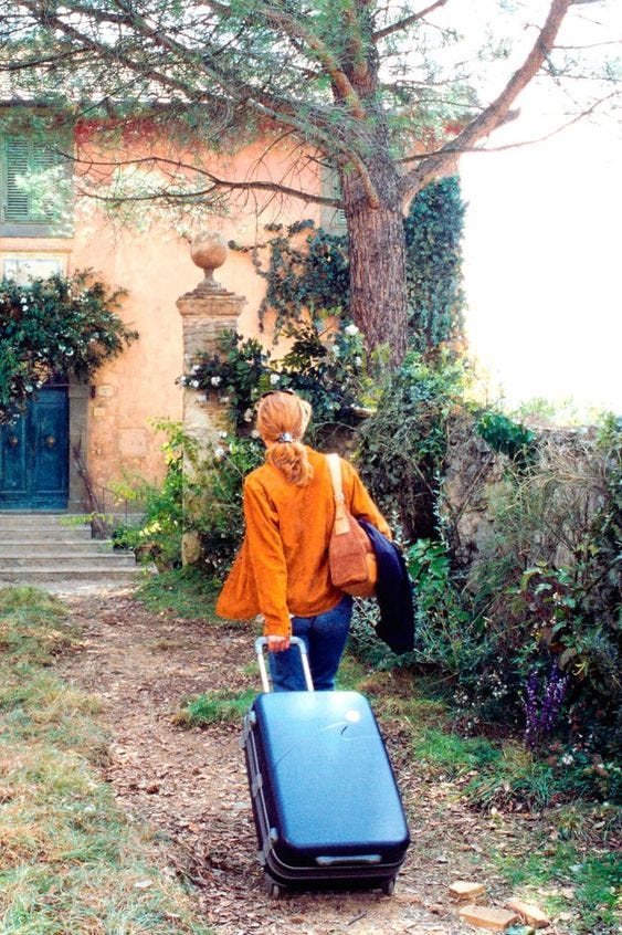 This may contain: a woman walking down a path with a blue piece of luggage in front of a house