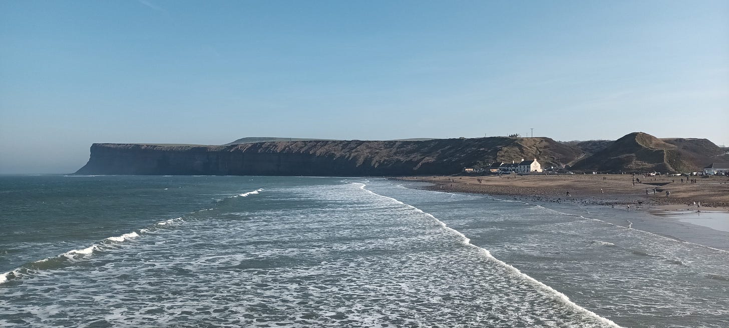 View from Saltburn Pier, waves coming into shore, cliffs in the distance, bright blue skies above