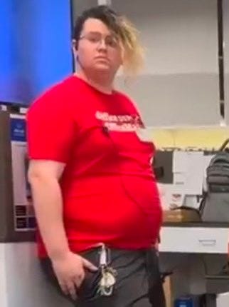 A person wearing a red T-shirt with text and a microphone clipped to it, standing in what appears to be an Office Depot store. The T-shirt has visible text reading "officedepot" on it. The person is also wearing dark pants and has a lanyard with a key or badge hanging from it. The background includes store shelves, a cash register, and a blue sign. A person wearing a red T-shirt with text and a microphone clipped to it, standing in what appears to be an Office Depot store. The T-shirt has visible text reading "officedepot" on it. The person is also wearing dark pants and has a lanyard with a key or badge hanging from it. The background includes store shelves, a cash register, and a blue sign.