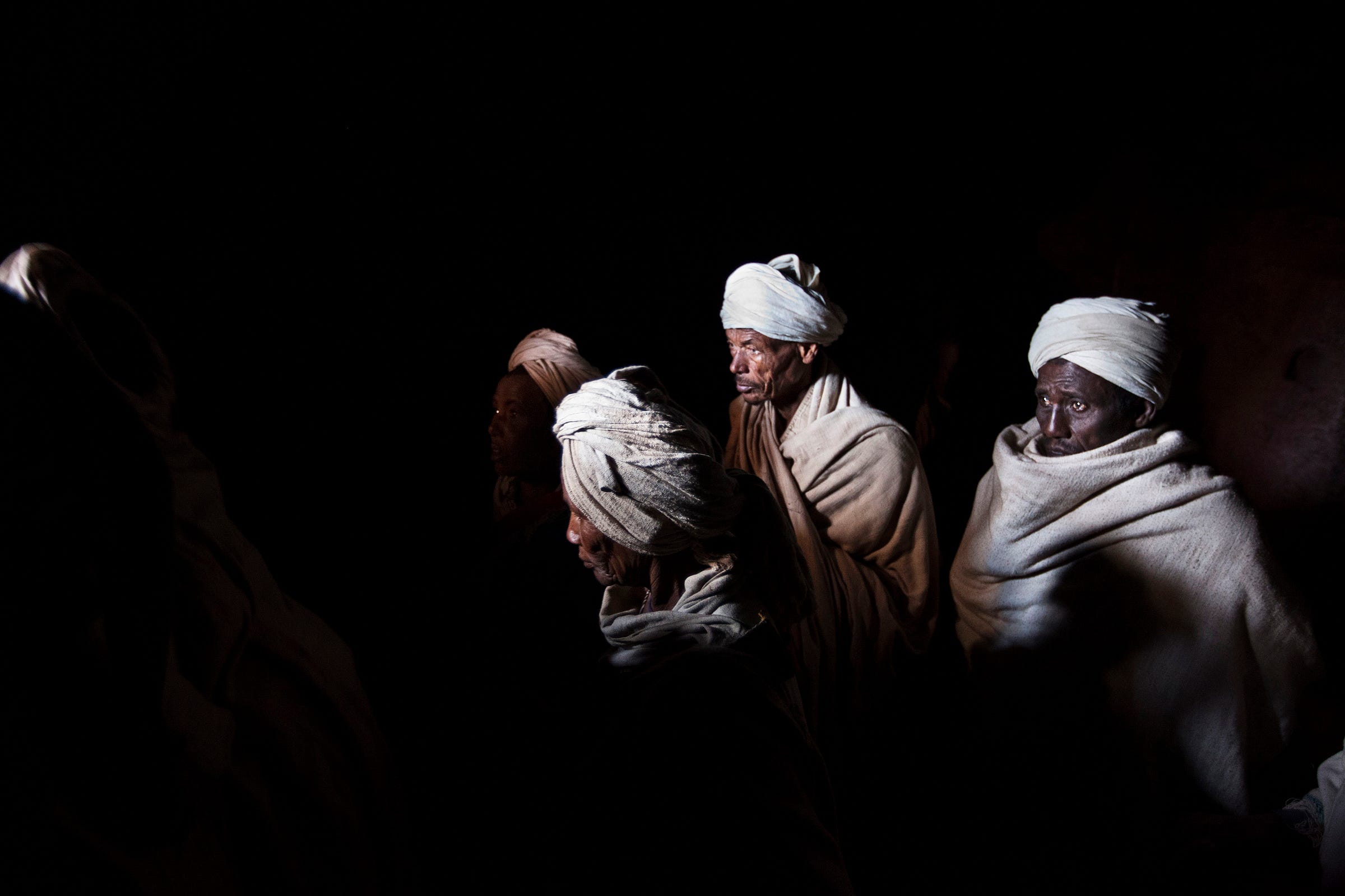 Four men wearing light-colored turbans and robes stand closely together, illuminated dramatically against a dark background, with their faces partially in shadow.