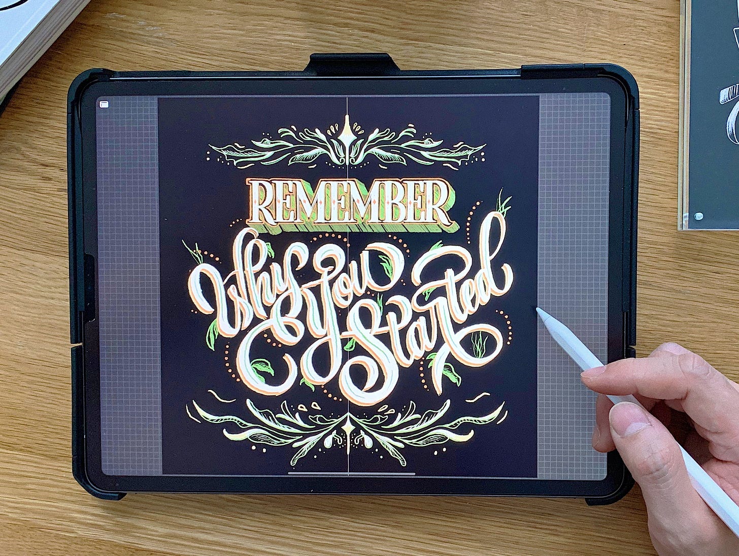 An iPad on a wooden desk with a hand holding an Apple Pencil beside it. On screen, a finished piece of hand lettering in gold and cream with green foliage, reading: REMEMBER WHY YOU STARTED. An iPad on a wooden desk with a hand holding an Apple Pencil beside it. On screen, a finished piece of hand lettering in gold and cream with green foliage, reading: REMEMBER WHY YOU STARTED.