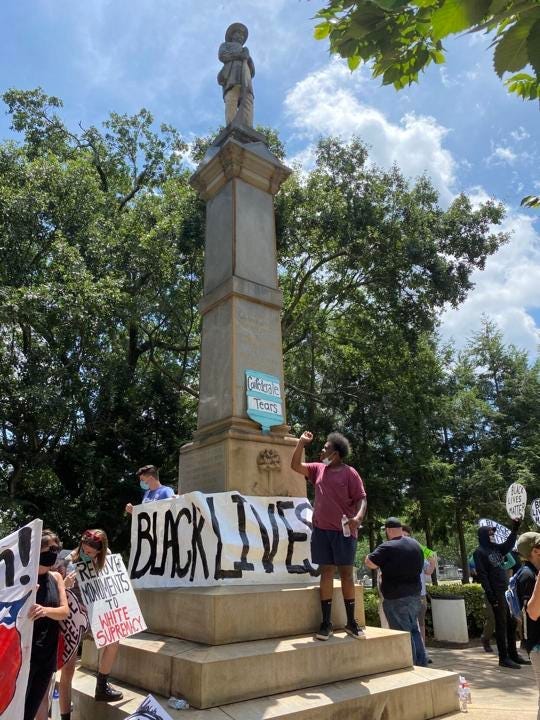 black man in burgundy shirt standing with fist raised standing in front of a confederate statue with a “Black Lives Matter” banner on it.